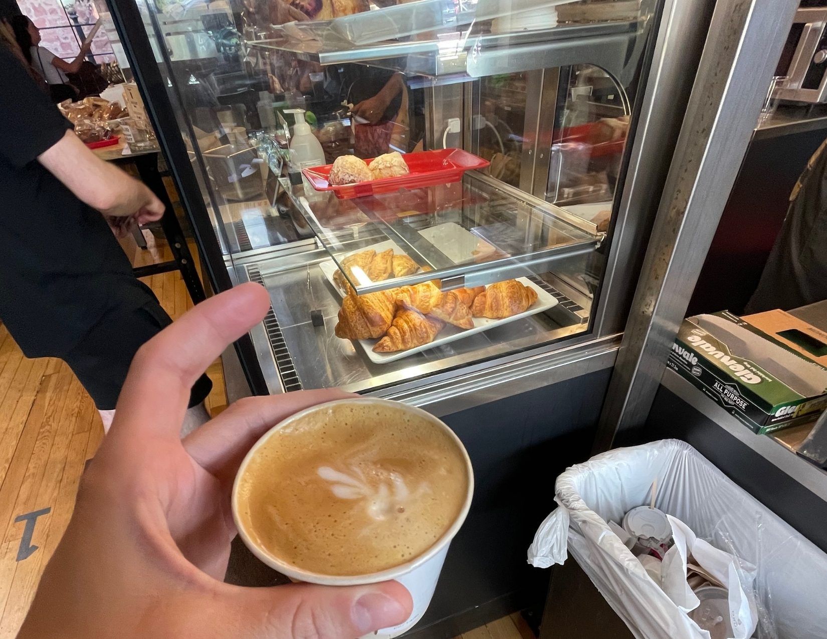 women's hand holding a latte with foam art at a coffee shop in Flagstaff, Arizona. in the background there is a case of pastries