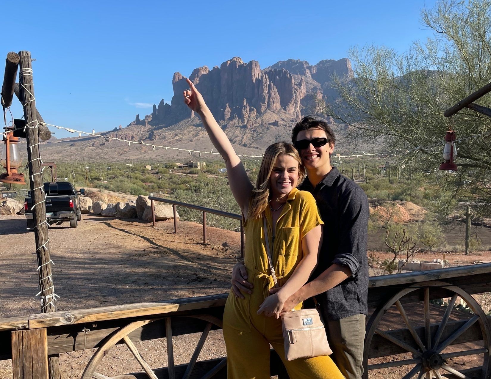 A young couple posing outside of a restaurant in Goldfield Ghost Town with purple and pink Superstition Mountains in the background.