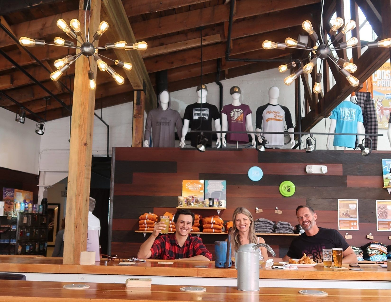 a young man holding up his beer, smiling next to two friends in a rustic yet modern brewery