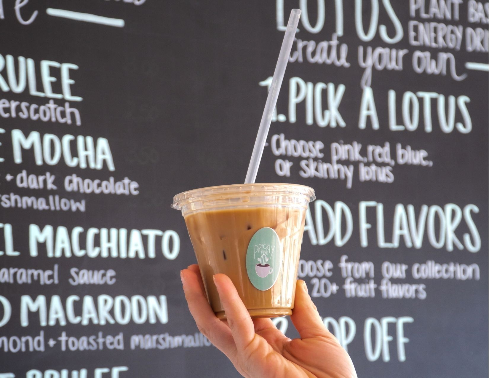 A hand holding an iced latte in front of a chalkboard menu in a coffee shop in Wickenburg, Arizona