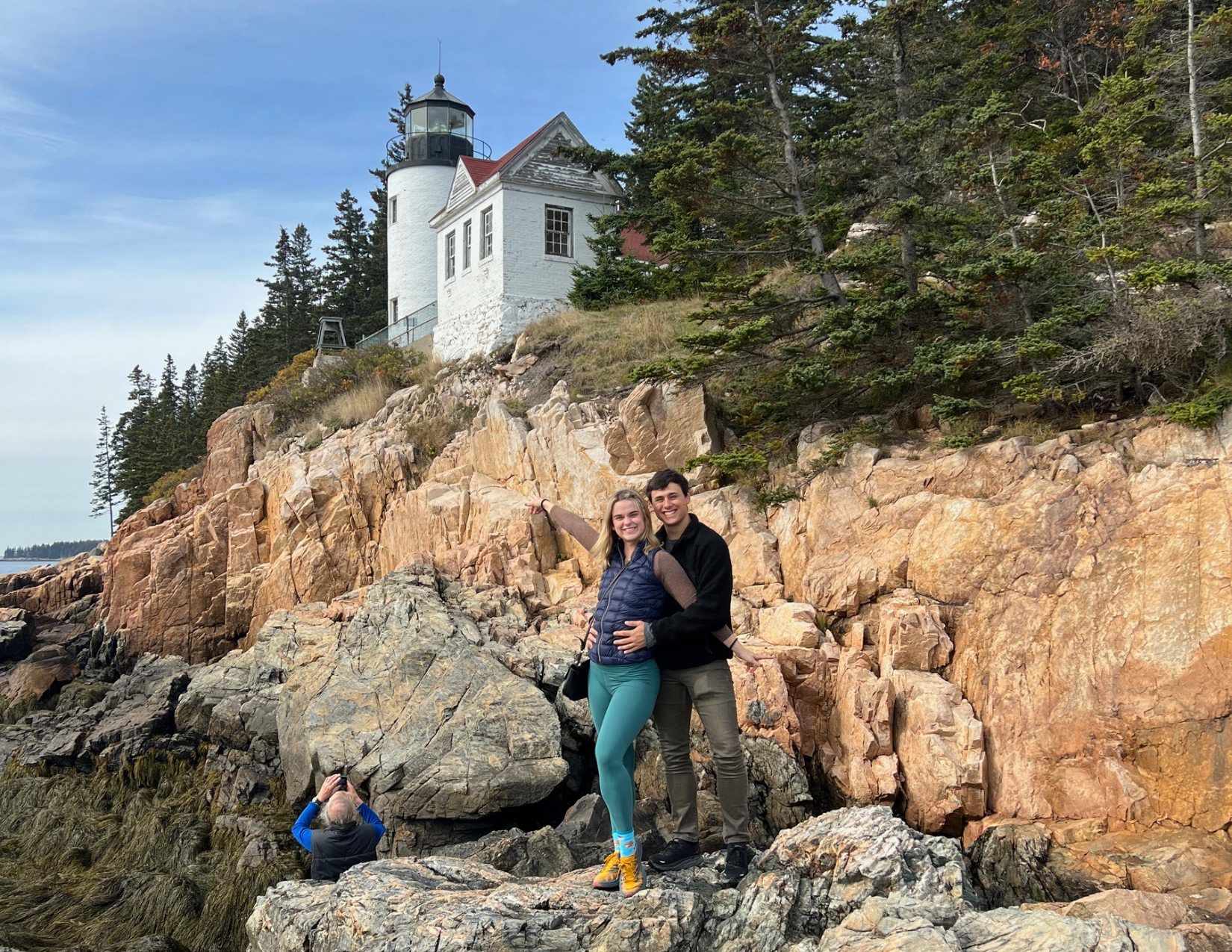 A couple smiling and standing on giant rocks in front of the Bass Harbor Head Lighthouse in Maine