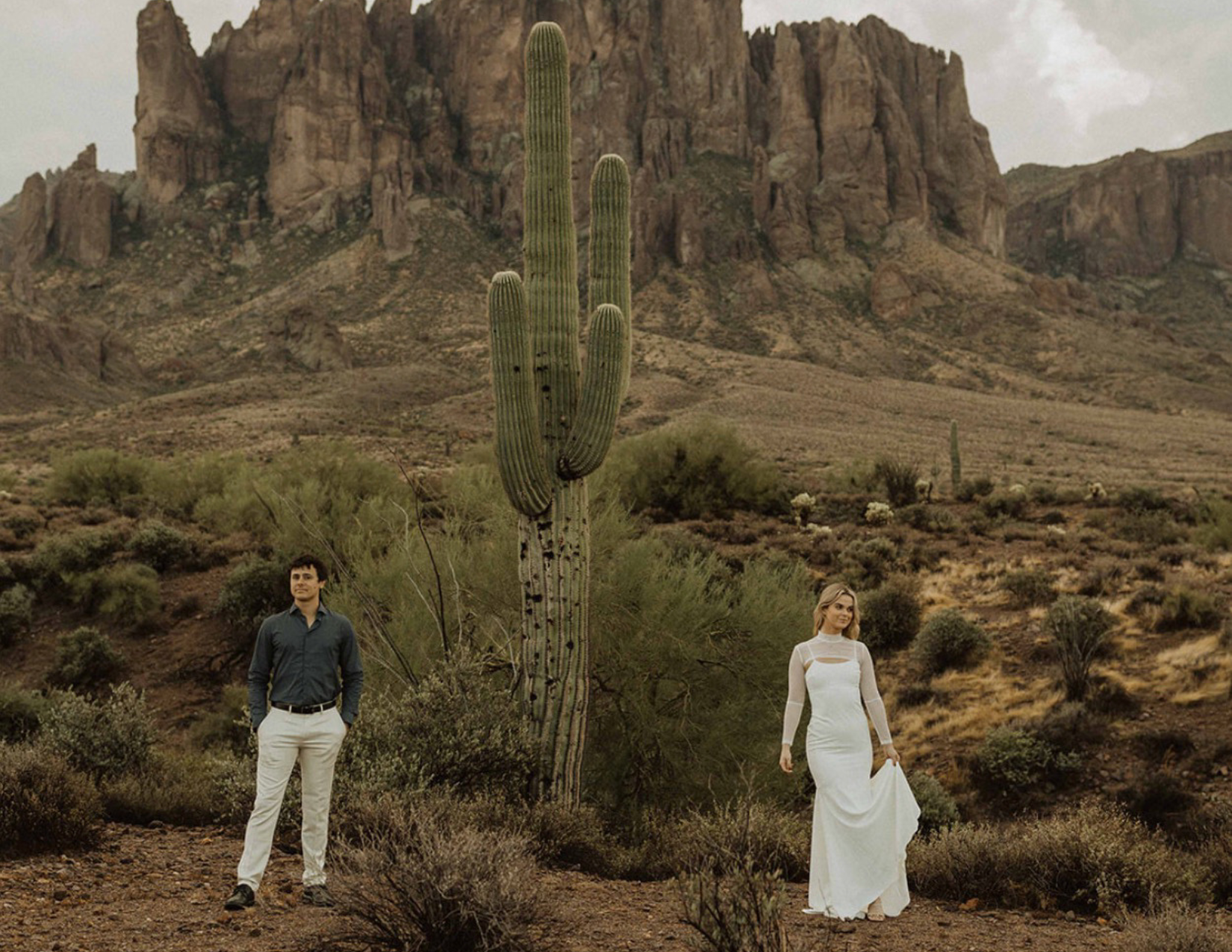 Man and woman stand on either side of large saguaro cactus in front of beautiful mountain scenery at Lost Dutchman State Park