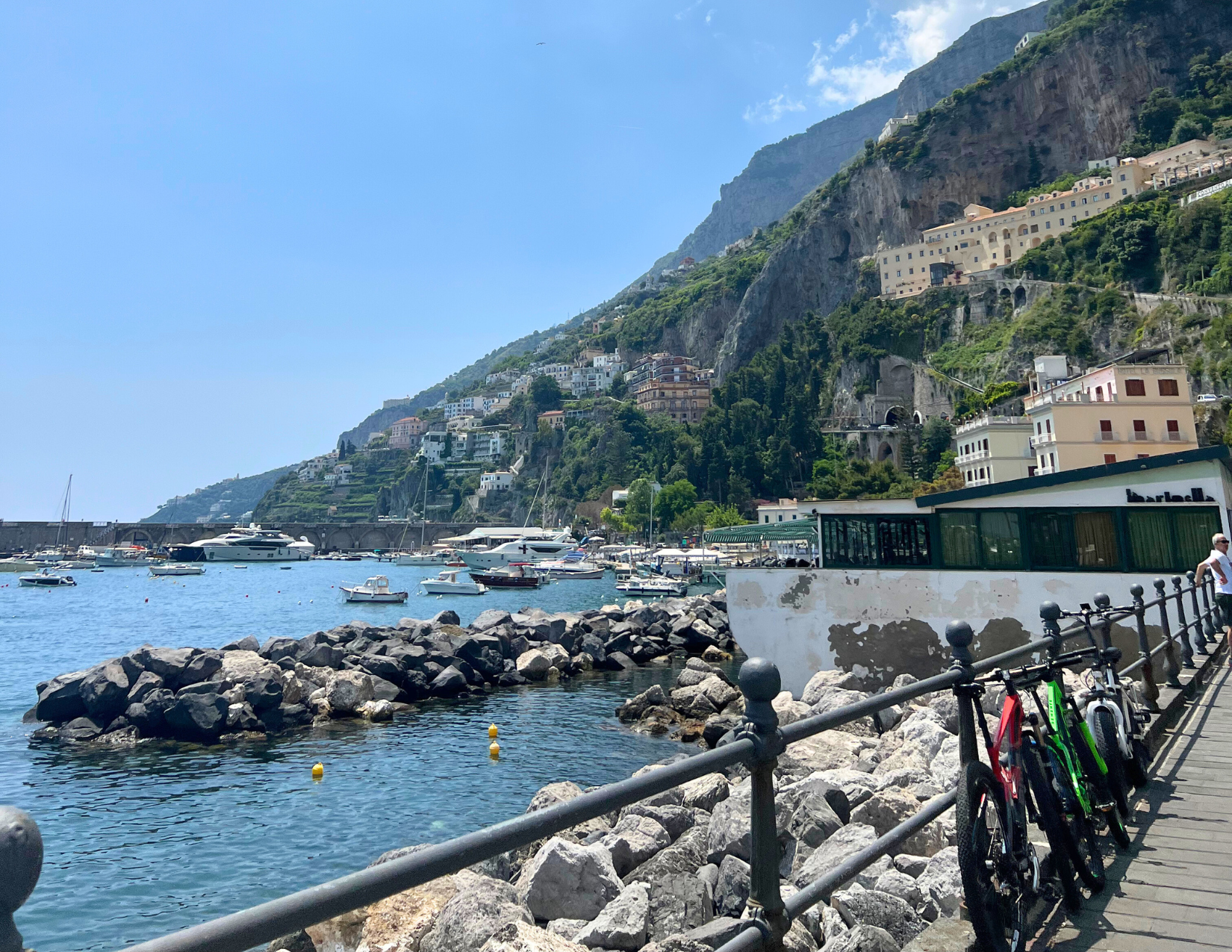 The boating dock in the town of Amalfi in Italy, with yachts docked in the marina and luscious green trees lining the hills. 