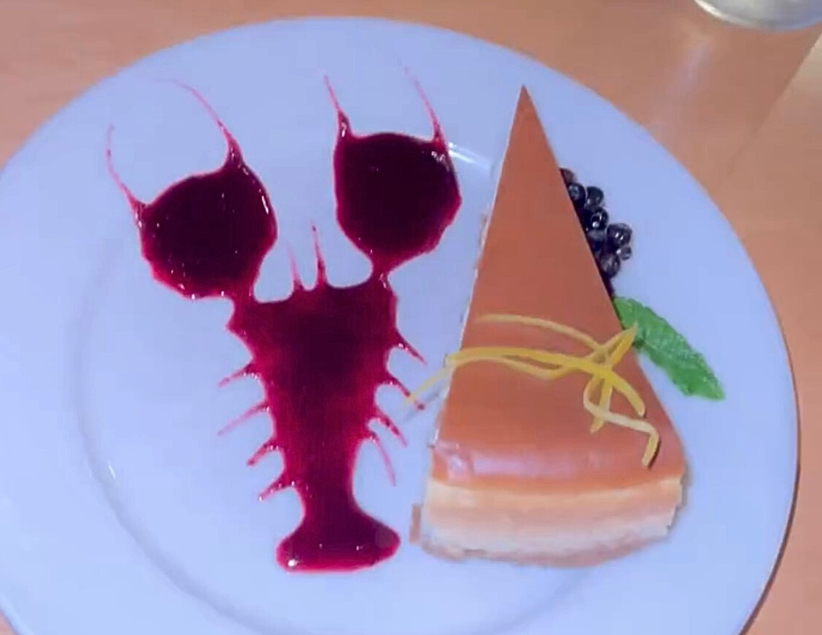 A slice cheesecake topped with lemon zest, sitting next to a lobster made out of blueberry juice