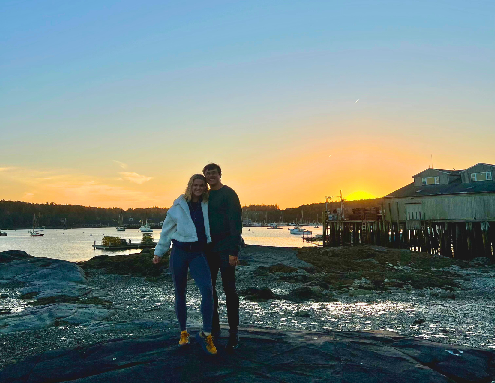 A couple smiling in front of a peaceful orange sunset on the harbor in Bar Harbor, Maine