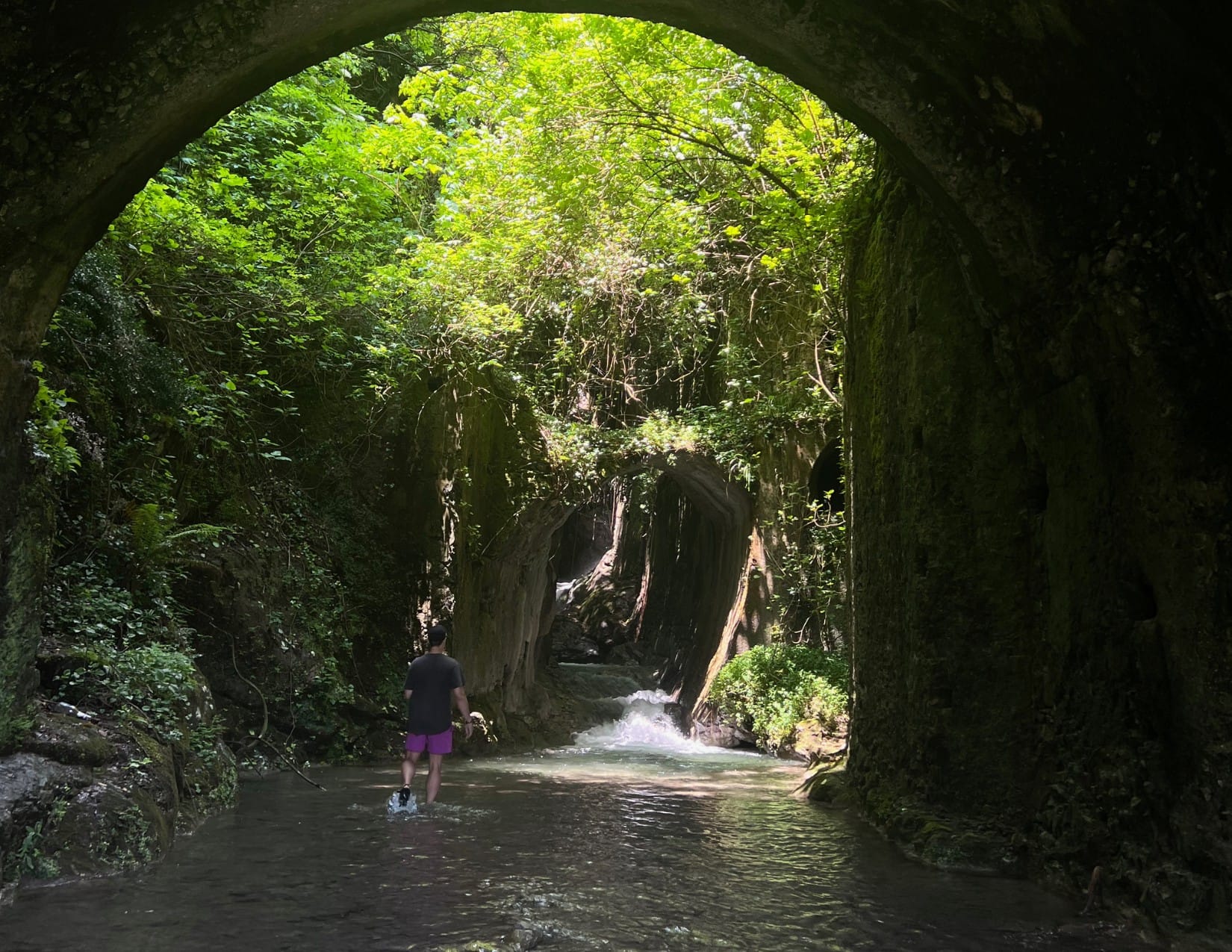 Man in purple shorts walking through water surrounded by overgrown green structures in Amalfi, Italy